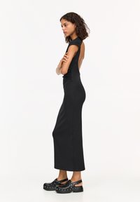 Schwarzes, ärmelloses Maxikleid mit offenem Rücken und gerafften Details. Modernes Design kombiniert mit schwarzen Clogs, die mit silbernen Hardware-Akzenten verziert sind.