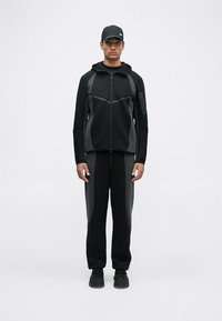 Completo sportivo nero composto da giacca con zip e pannelli grigi a contrasto e pantaloni tapered coordinati. Include un cappellino e sneakers.