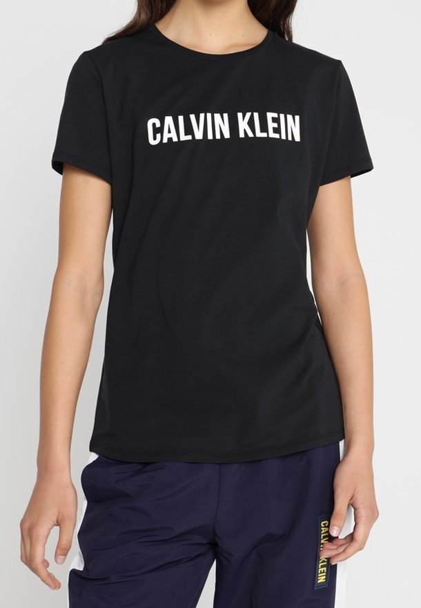 Mujer vistiendo una camiseta negra de Calvin Klein y pantalones azul marino de Calvin Klein con parche del logo amarillo en las franjas laterales blancas.