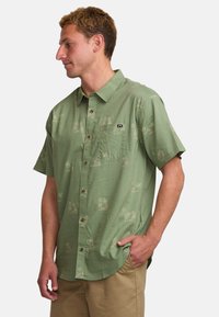 Camicia a maniche corte con bottoni in tessuto verde chiaro, caratterizzata da un sottile motivo di palme e una tasca sul petto, con bottoni marroni e una vestibilità comoda.