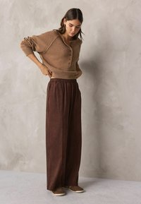 Brauner Strickpullover mit einem Knopfverschluss am Ausschnitt, kombiniert mit lockeren, dunkelbraunen plissierten Hosen. Das Model steht auf einem hellgrauen Boden.