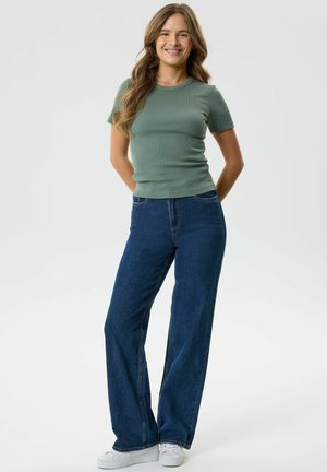 Joven mujer con cabello largo y ondulado que lleva una camiseta verde ajustada, vaqueros azules de pierna ancha y zapatillas blancas, de pie con las manos detrás de la espalda.