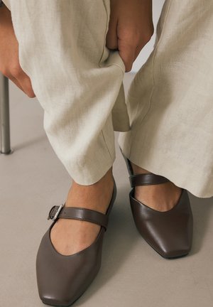Persona che aggiusta pantaloni beige a gamba larga, indossando scarpe Mary Jane marrone scuro con cinturino a fibbia, in piedi su un pavimento chiaro.