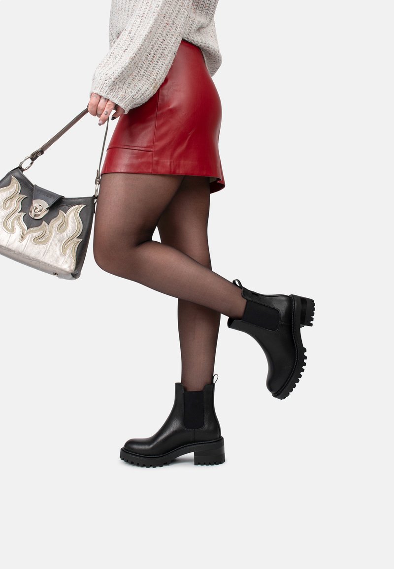 Botas negras de cuero hasta el tobillo con paneles laterales elásticos, suelas de goma gruesa y un acabado texturizado, combinadas con una falda roja de cuero y medias transparentes.