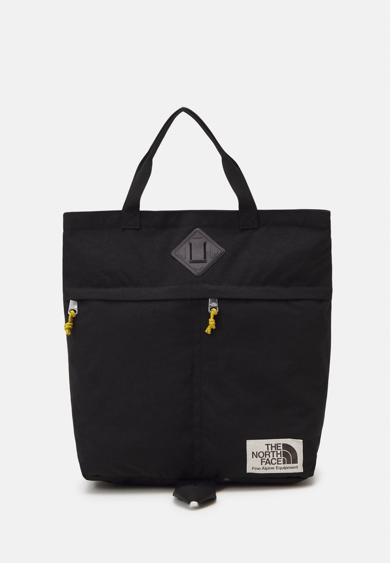 Sac cabas noir avec deux poignées, poche zippée à l’avant avec tirettes jaunes, et écusson du logo The North Face dans le coin inférieur droit.
