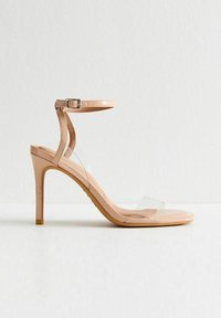 Beige högklackad sandalett med en klar plastrem och en ankelspänne. Har en slank form, slät yta och en tunn stilettklack.