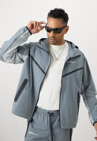 Ανοιχτό γκρι αδιάβροχο με κουκούλα, μαύρες λεπτομέρειες, τσεπάκια με φερμουάρ. Συνδυασμένο με γκρι σορτσάκι και λευκό μπλουζάκι. Φοριούνται γυαλιά ηλίου.