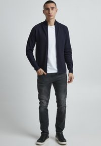 Tmavomodrá zipová bunda s rebrovanou textúrou, nosená na bielom tričku. Šedé slim-fit džínsy a čierne tenisky dopĺňajú tento outfit.