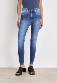 Jeans ajustados de color azul con un lavado medio, cinco bolsillos y un ligero desgaste, combinados con sandalias negras de tacón bloque.