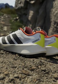 Chaussures de trail Adidas Terrex Agravic 4 blanches, noires, orange et jaunes reposant sur un sol rocheux avec en arrière-plan des rochers naturels flous.