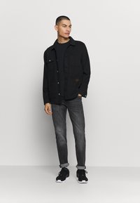 Homem em pé contra um fundo simples, vestindo uma jaqueta preta, uma camisa preta, jeans cinza arregaçados e sapatilhas pretas.