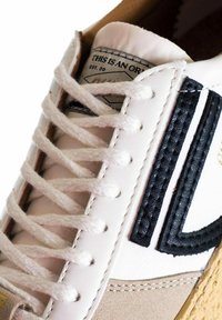 Sneaker bianco con un logo nero testurizzato, lacci crema e un accento in suede beige. Presenta un dettaglio cucito e un'etichetta sulla linguetta.