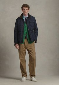 Μπλε σκούρο μπουφάν με καφέ γιακά, πράσινη ζακέτα, καφέ παντελόνι με σχέδια και λευκά αθλητικά παπούτσια. Το μοντέλο στέκεται μπροστά σε ουδέτερο φόντο.