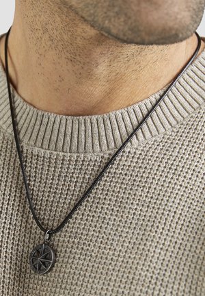 Ledern halskette mit einem runden, geprägten Kompassanhänger, der über einem strukturierten, hellgrauen Strickpullover mit geripptem Ausschnitt hängt.