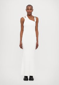 White ribbed knit dress with asymmetrical neckline, single strap with buckle detail, and floor-length cut. Worn with black boots.