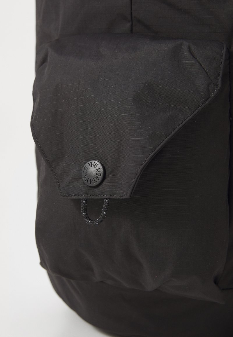Sac à dos noir avec un tissu texturé, un rabat avec une fermeture à bouton et un détail à cordon. Le design est élégant et minimaliste.