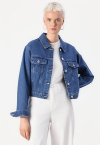 Denim jakna v srednje modri barvi s skrajšanim dizajnom, sprednjimi žepi, srebrnimi dodatki in gumbom za zapiranje. Nošena preko bele majice in svetlih hlač.