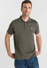 Camisa polo verde oliva de manga corta, con cuello acanalado, tapeta de tres botones y un pequeño parche con el logo; combinada con jeans de mezclilla azul.