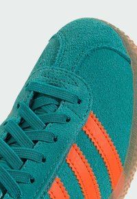 Teal suede sneaker with orange zigzag stripes, textured upper, and green laces. Translucent rubber sole. Close-up view of design elements.
