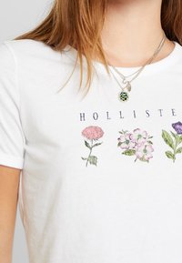 White cotton t-shirt featuring embroidered flowers in pink, purple, and green. The word "HOLLISTER" is printed above the design.