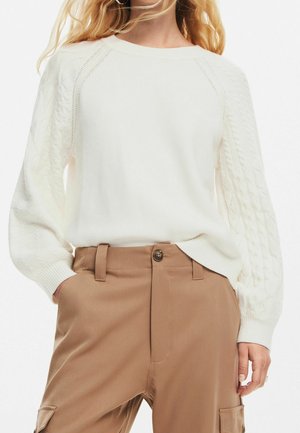 Mujer con suéter blanco de punto de ochos con mangas abullonadas y pantalones marrón claro de tiro alto con las manos en los bolsillos.