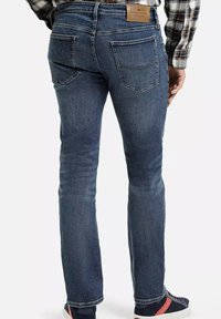 Mężczyzna w ciemnoniebieskich, dopasowanych dżinsach i koszuli w kratę, pokazany od pasa w dół, stojący na tle białym.