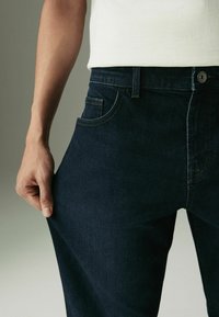 Tamsiai mėlynos džinsinės kelnes su klasikiniu kirpimu, turinčios kišenę priekyje, diržo kilpas ir matomą metalinį mygtuką ant liemens.