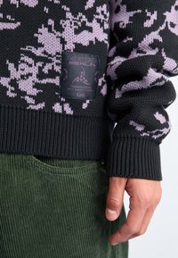 Pulover negru cu un model de camuflaj roz, textură tricotată și manșete cu nervuri. Prezintă o etichetă neagră cu logo și text. Purtați peste pantaloni din catifea verde.