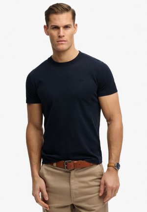 Uomo che indossa una maglietta a maniche corte blu navy, pantaloni beige con cintura marrone e orologio da polso, in piedi contro uno sfondo bianco semplice.