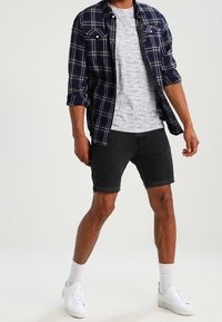 Chemise en flanelle à carreaux marine sur un t-shirt gris, associée à un short en denim noir et des baskets blanches. Chaussettes blanches visibles au-dessus des chaussures.
