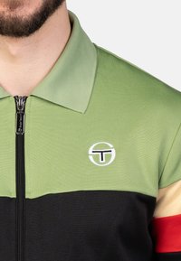 Groene en zwarte rits polo shirt met een logo, contrasterende panelen en een gestructureerde kraag. Accenten omvatten tinten rood en crème op de mouwen.