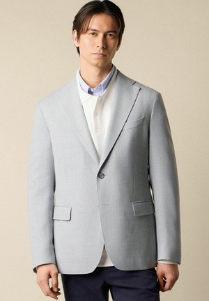 Man draagt een lichtgrijze blazer over een witte polo en gestreept overhemd, voor een effen beige achtergrond, kijkt recht vooruit met een neutrale uitdrukking.