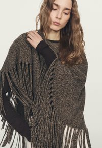 Poncho marrone lavorato a maglia con frange, con una vestibilità ampia e un motivo testurizzato. Indossato sopra una maglia a manica lunga scura.