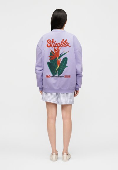 Άτομο που φοράει μωβ φούτερ με μεγάλο κόκκινο και πράσινο λουλούδι παραδείσου και το κείμενο "Stieglitz" στην πλάτη, σε συνδυασμό με κοντά παντελόνια καρό.