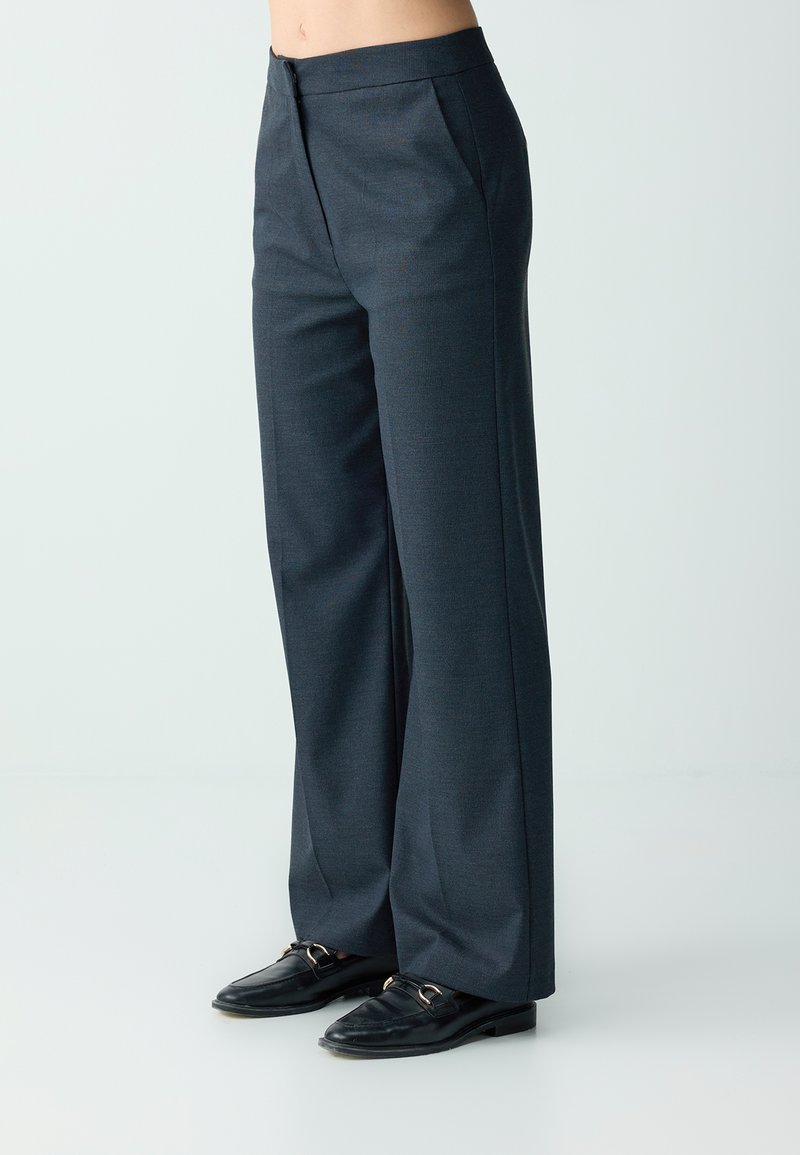 Personne portant un pantalon gris à jambes larges avec poches latérales et des mocassins noirs, debout sur un fond clair uni.