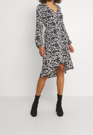 Frau trägt ein schwarz-weißes Blumenmuster-Wickelkleid mit langen Ärmeln, asymmetrischem Saum und schwarzen Knöchelstiefeln, steht vor einem schlichten Hintergrund.
