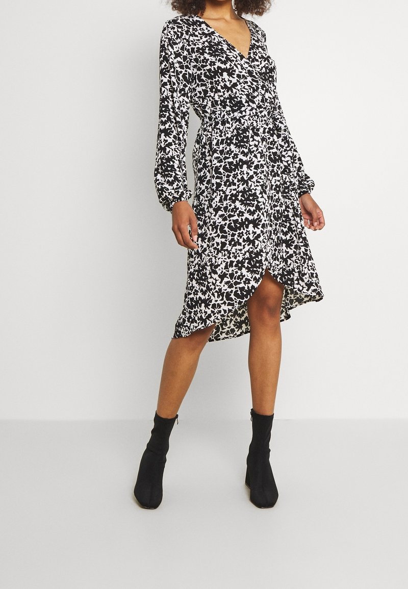 Femme portant une robe portefeuille noire et blanche à motif floral avec manches longues, un ourlet asymétrique et des bottines noires, debout devant un fond uni.