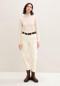 Suéter de cuello alto crema combinado con pantalones de cintura alta en color blanco roto y botas de tobillo marrones con suelas negras gruesas. Cinturón trenzado marrón como acento.