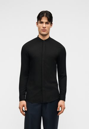 Bărbat purtând o cămașă neagră, mulată, cu guler mandarin și nasturi ascunși, și pantaloni întunecați cu dungi subțiri, stând în fața unui fundal simplu.