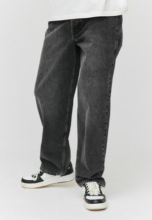 Jean en denim noir avec une coupe ample, présentant une finition subtilement texturée et une couture contrastante, assortis à des baskets noires et blanches.