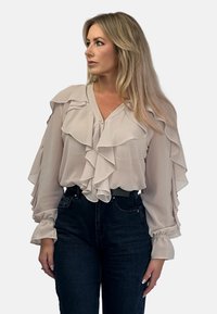 Jasno beżowa bluzka z falbanami na rękawach i przodzie, wykonana z przejrzystego materiału, zestawiona z ciemnoniebieskimi dżinsami.