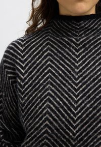 Črno-beli pulover s črtastim vzorcem chevron, z visokim ovratnikom in mehko teksturo, z drobnimi detajli in prijetnim videzom.