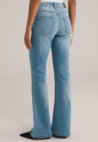Blaue, ausgestellte Jeans aus Denim mit einem verwaschenen Finish. Enthält zwei Gesäßtaschen und eine hohe Taille. Schwarze, spitze Ballerinas runden den Look ab.