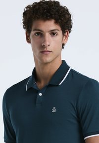 Dunkelblaues Poloshirt mit einem weißen Kragen, einer drei-Knopf-Leiste und einem kleinen Pinguin-Logo auf der linken Brust. Glatte Stofftextur.