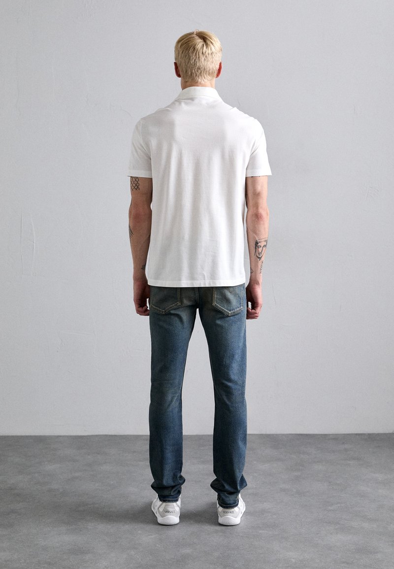 Man med kort blont hår som bär en vit polo, blå jeans och vita sneakers, står med händerna vid sidorna vänd bort.