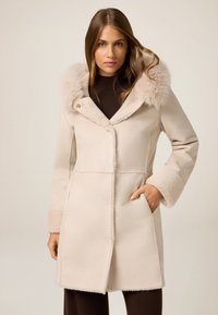 Cappotto beige con colletto e polsini in pelliccia sintetica, caratterizzato da una chiusura con bottoni, tasche laterali e un tessuto morbido e testurizzato.