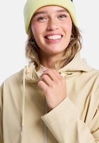Beige Regenjacke mit einem Reißverschluss und Kapuze mit Kordelzug, kombiniert mit einer hellgelben gerippten Mütze. Freundliches Lächeln und natürliche Haarstruktur sichtbar.