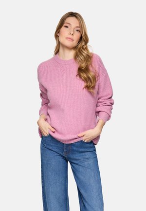 Mujer con cabello largo y ondulado, vestida con un suéter rosa acanalado y vaqueros azules, de pie con las manos en los bolsillos frente a un fondo sencillo.