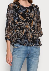 Sort gennemsigtig bluse med blå og orange blomsterprint, rund halsudskæring, samlet talje og trekvartærmer, parret med lyseblå jeans.