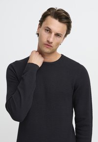 Pulover negru tricotat cu un model texturat, guler rotund și manșete cu blăniță, purtat de un model cu o expresie neutră.